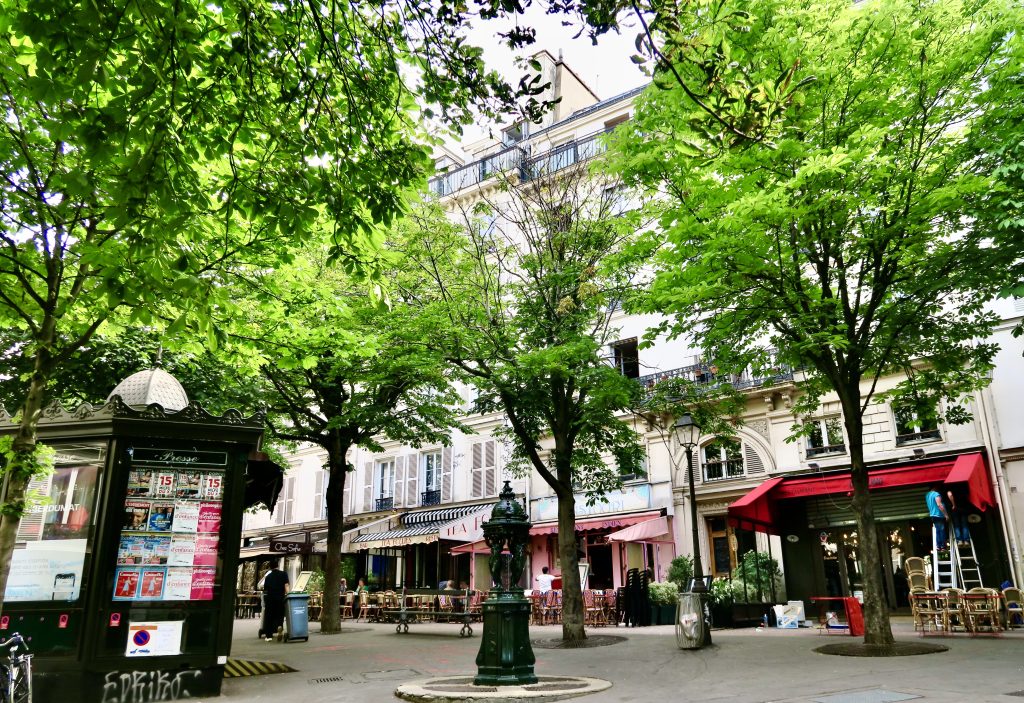 Photo en couleur de la place Touldouze à Paris 9ème avec fontaine, kiosque, terrasses de cafés et arbres.