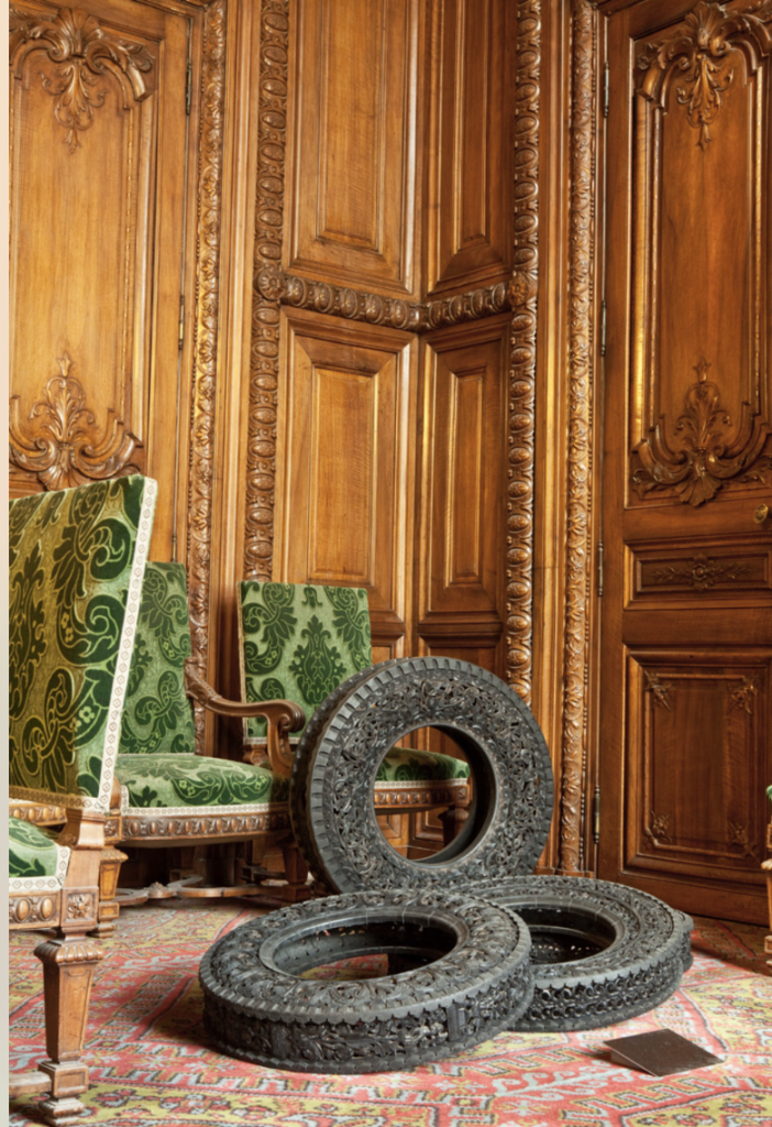 Trois pneus en caoutchouc entièrement sculptés posés sur le sol recouvert d'un tapis, dans une pièce avec murs de lambris en bois, et un fauteuil en velours vert.