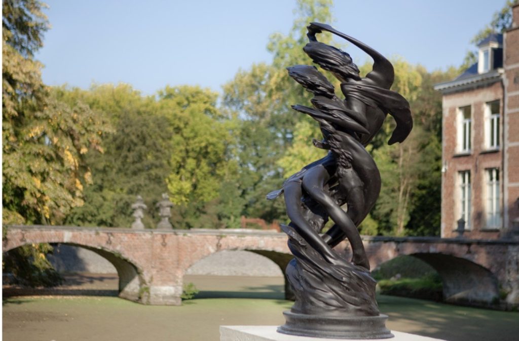 Sculpture en bronze d'un couple formant une spirale ascendante, avec en arrière-plan un pont en brique, une maison sur la droite, et des arbres au fond.