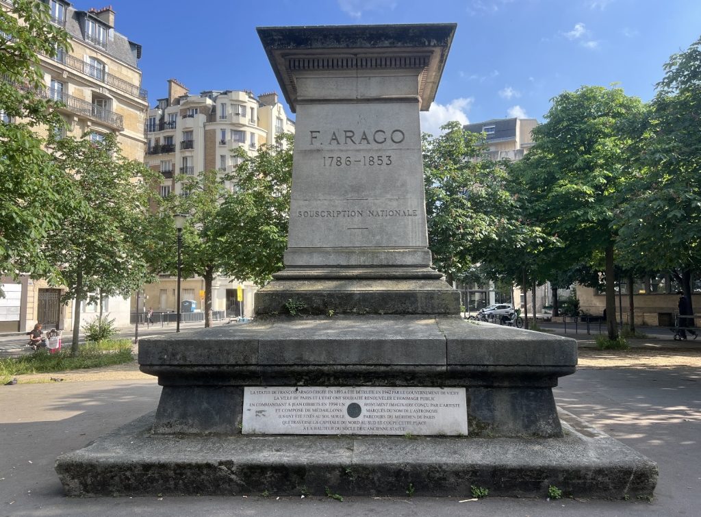 Photo d'un socle en pierre avec une inscripition gravée F.ARAGO 1786-1853. Une plaque de marbre portant un texte est posée au pied du socle. Des arbres et immeubles en briques sont présents à l'arrière-plan.