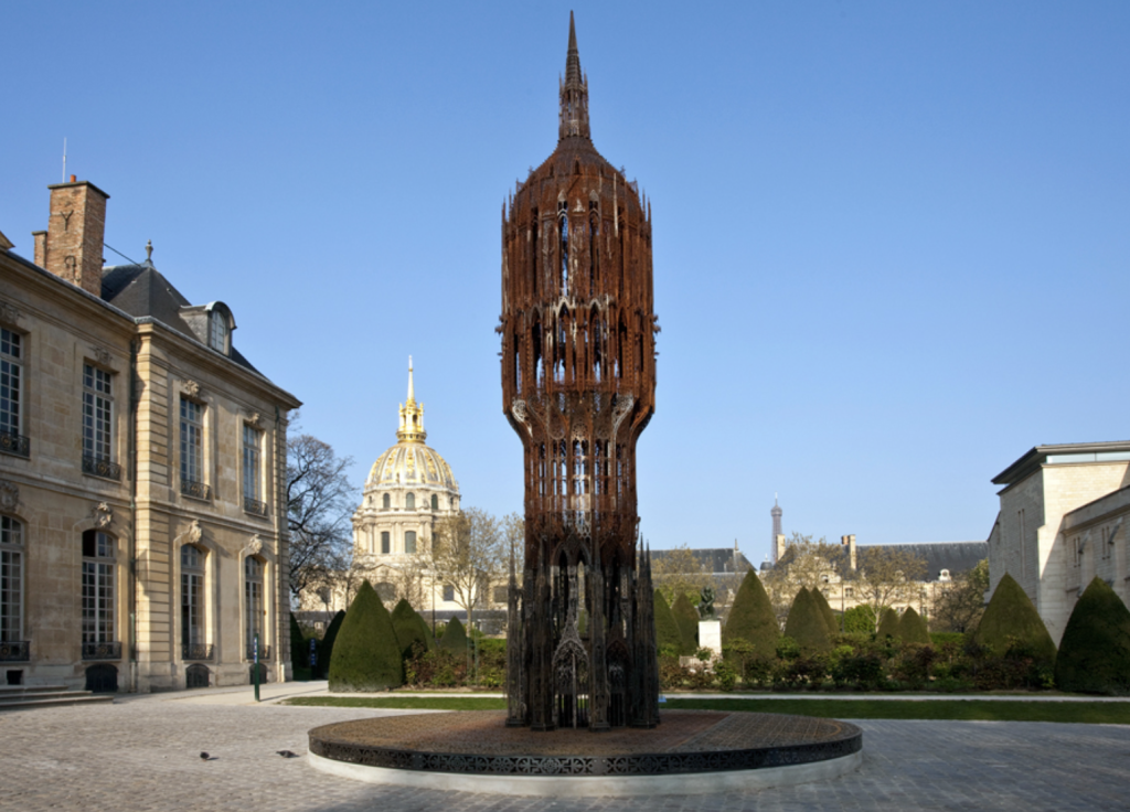 Photo d'une tour en acier ajouré , dans la cour d'entrée du Musée Rodin, à Paris, avec à l'arrière plan le dôme des Invalides (à gauche) et la tour Eiffel (à droite).