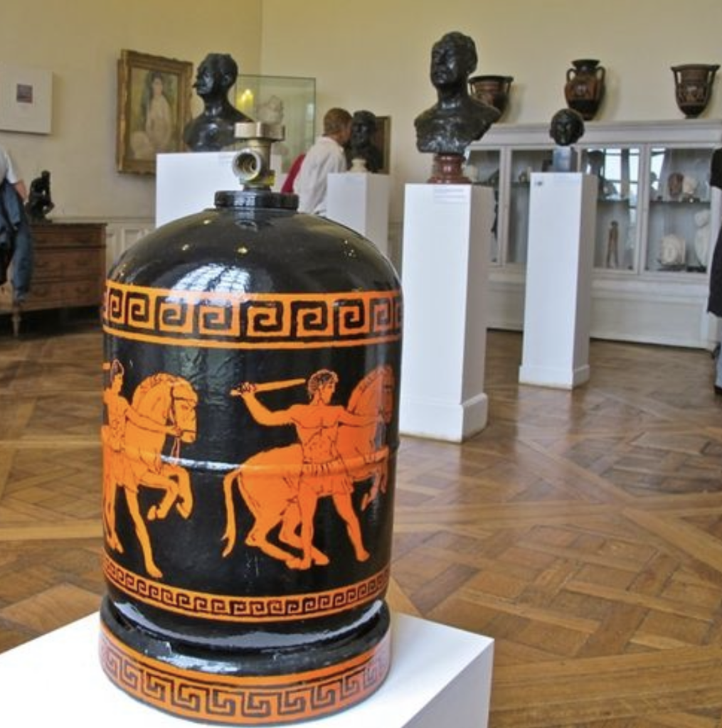 Photo d'une bonbonne de gaz, entièrement couverte d'un décor peint émaillé de motifs de cavaliers grecs se détachant en orange sur fond noir. Elle est posée sur un socle, dans une salle de musée, avec des bustes en bronze à l'arrière-plan.