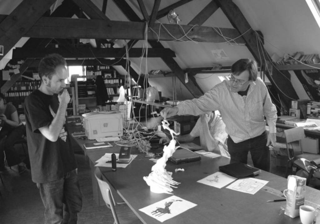 Photo en noir et blanc de deux hommes debout autour d'une table sur laquelle sont posés des maquettes et des dessins. L'atelier a une structure porteuse en bois. Tout autour, de nombreuses étagères, une imprimante, et autres instruments de travail.