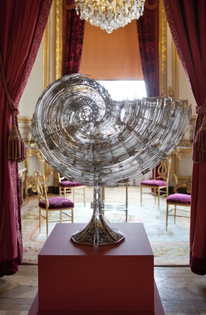 Photo en couleur avec au premier plan une sculpture en acier d'un très gros coquillage, posée sur un socle rouge, et en arrière-plan un salon avec des rideaux et des sièges en velours rouge, un tapis et un lustre.