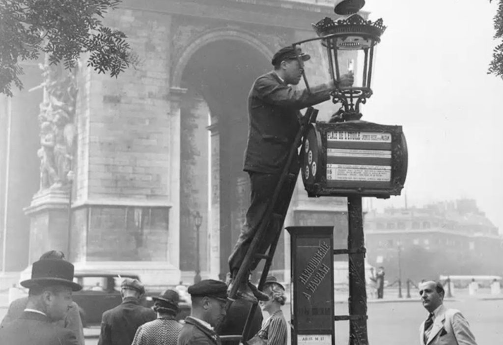 Photo noir et blanc d'un homme en uniforme en haut d'une échelle, allumant un réverbère. Autour de lui, de nombreux passants. A l'arrière-plan, l'Arc de Triomphe, Place Charles de Gaulle à Paris.