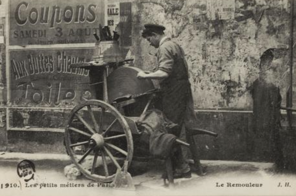 Photo noir et blanc d'un rémouleur actionnant une meule en pierre fixée à une charrette, devant une façade dégradée partiellement couverte de vieilles affiches.