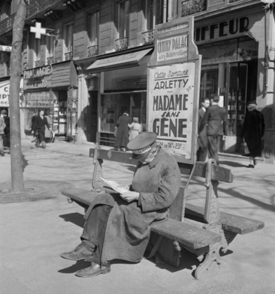 Photo noir et blanc d'un homme coiffé d'une casquette, lisant un journal sur un blanc dans une rue parisienne. Il porte dans le dos un grand panneau avec l'inscription "Arletty, Madame Sans Gêne.