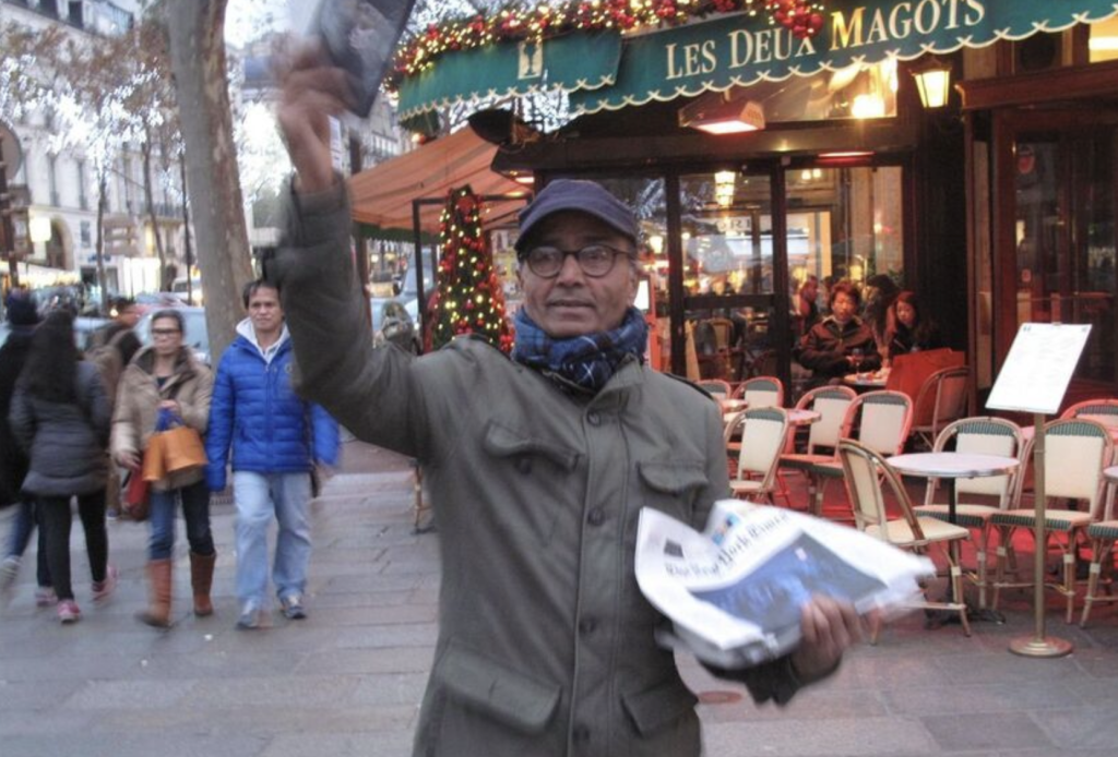 Photo en couleur d'un homme coiffé d'une casquette, brandissant un journal de la main droite et tenant les autres journaux de son bras gauche. En arrière-plan, la terrasse du café des Deux Magots, à Paris, ainsi que des passants.