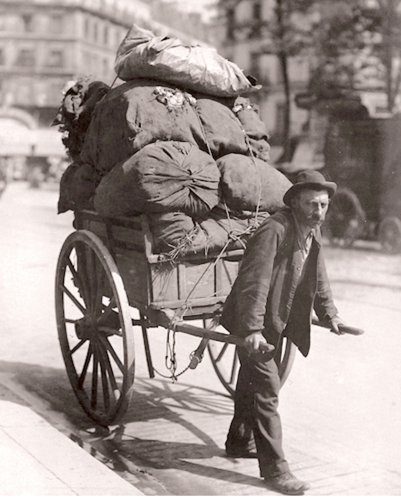 Photo en noir et blanc d'un homme tirant une charrette pleine de balluchons maintenus par une ficelle, dans une rue de Paris.