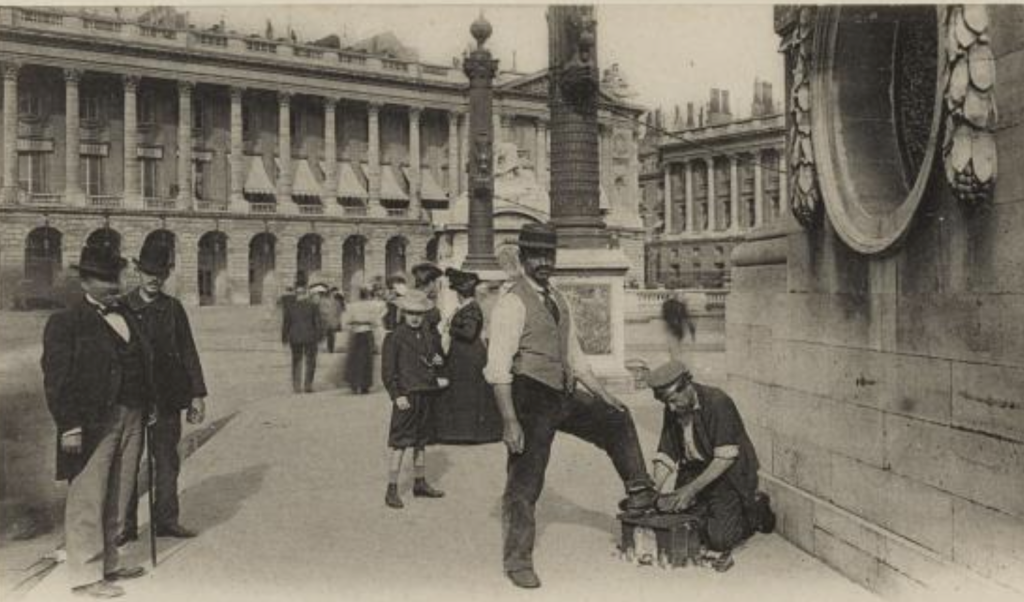 Photo en noir et blanc d'un homme agenouillé, cirant les bottes d'un client. Deux hommes sur la gauche sont de trois-quart, et à l'arrière-plan un jeune garçon et d'autres femmes et hommes s'éloignent. La scène se passe sur la Place de la Concorde à Paris.